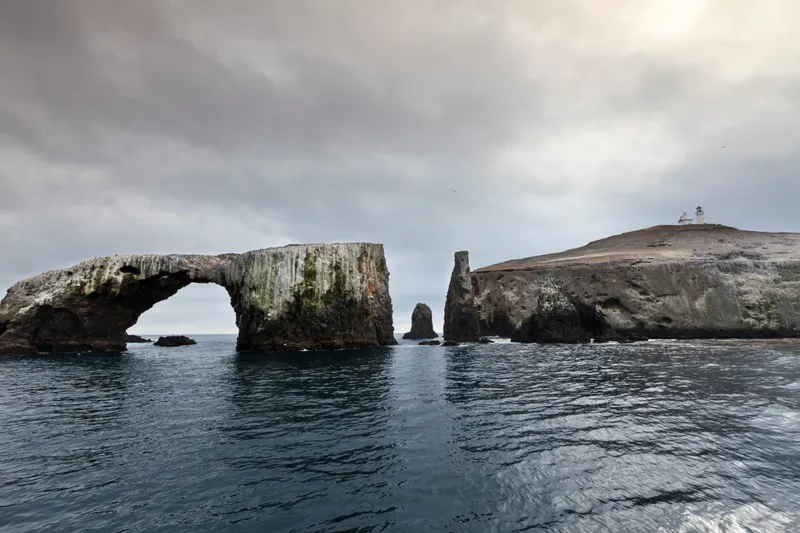 Steep dark volcanic rock has been eroded into a 40-foot-high arch that is surrounded by water. Behin