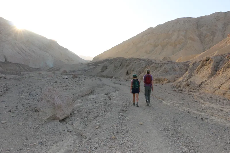 Two hikers walk up a wash with their backs to the camera, while the sun rises over the canyon wall.