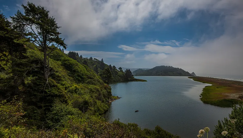 A calm lagoon with blue water. Hills with trees on the left, a spit of sand and ocean to the right.