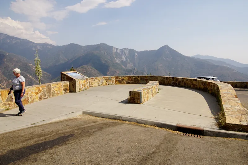 A curving rock wall sits above an asphalt highway. In the distance tall crags of rock can be seen